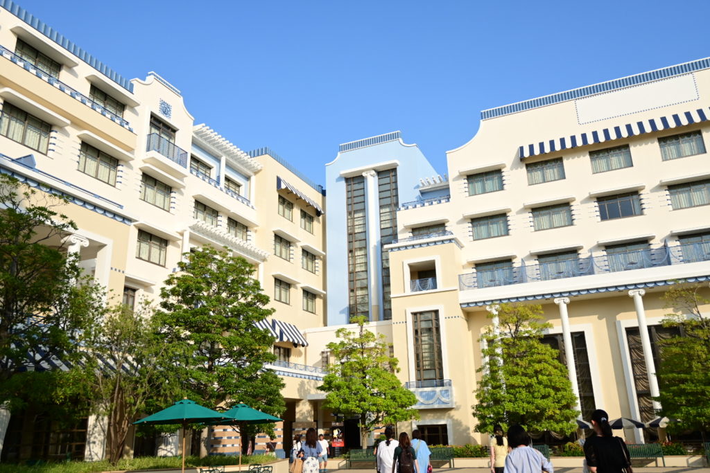 劇場に向かう途中にあるディズニーアンバサダーホテルの外観。奥には劇団四季『美女と野獣』の看板があり、青空の下、多くの人が看板を横目に劇場方面へと歩く様子。