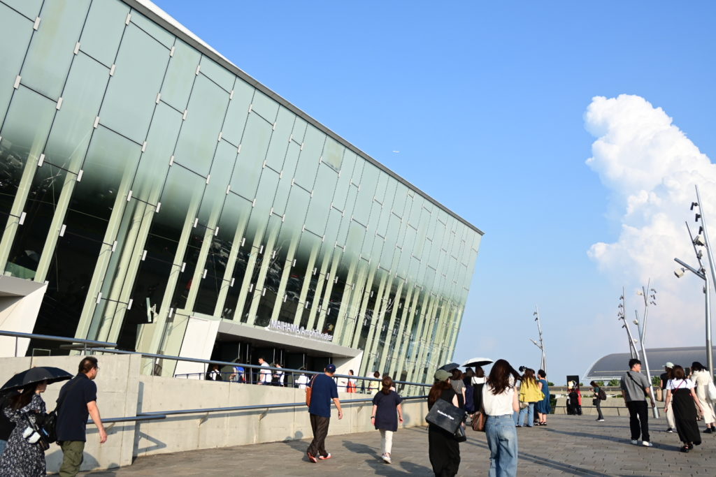 ガラス張りの舞浜アンフィシアターの外観と、入り口に向かって歩く観客たち。期待に満ちた開演前のひととき。パキッとした青空に、ガラス張りのシアターが映える。