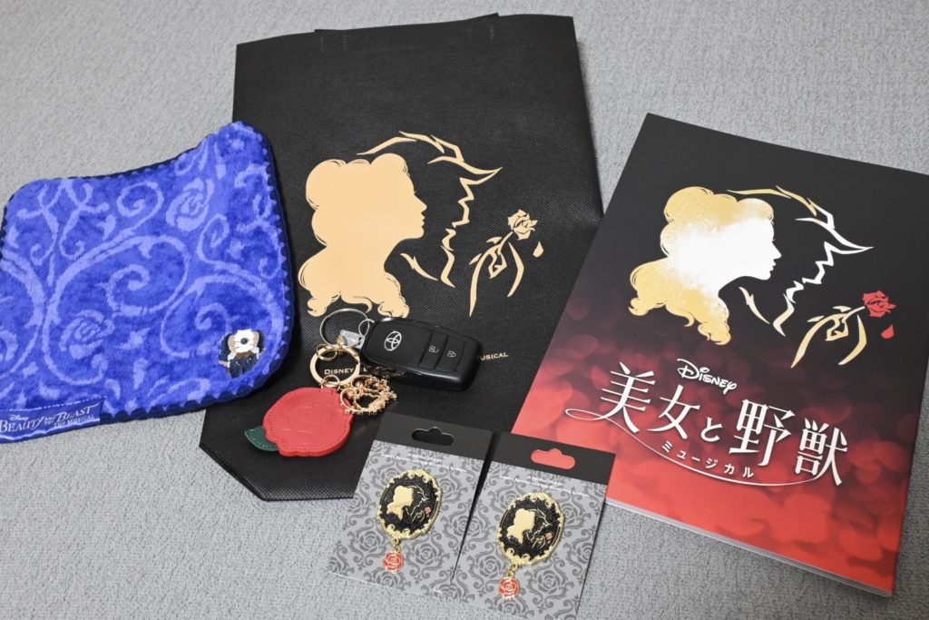 美女と野獣の観劇グッズを自宅で撮影。パンフレットやピンバッチ、バラのチャーム、青いハンドタオル、お買い物バッグが並ぶ。