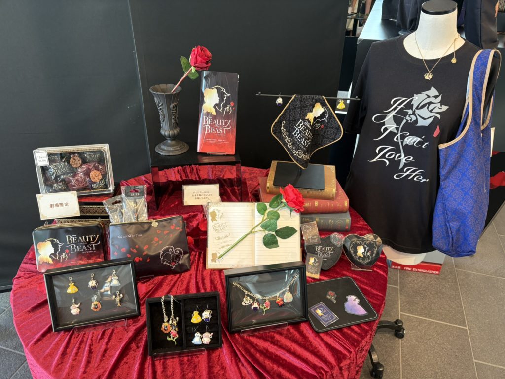 劇団四季『美女と野獣』のグッズ展示コーナー。お菓子、チャーム、Tシャツ、ハンドタオルなど多彩なアイテムが赤いテーブルクロスの上に美しく並べられている様子。
