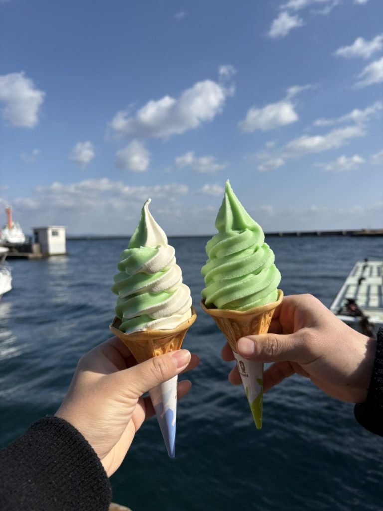 別府湾を背景に、コーンに盛られた黄緑色のかぼすソフトクリームを手に持つ様子